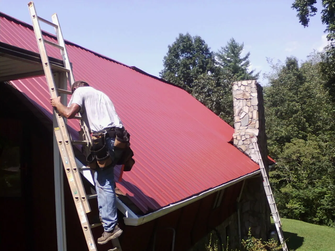 Roofer on a ladder beside a red metal roof and stone chimney — Soffit & Fascia Repair by Roofing Brothers in Monmouth Junction